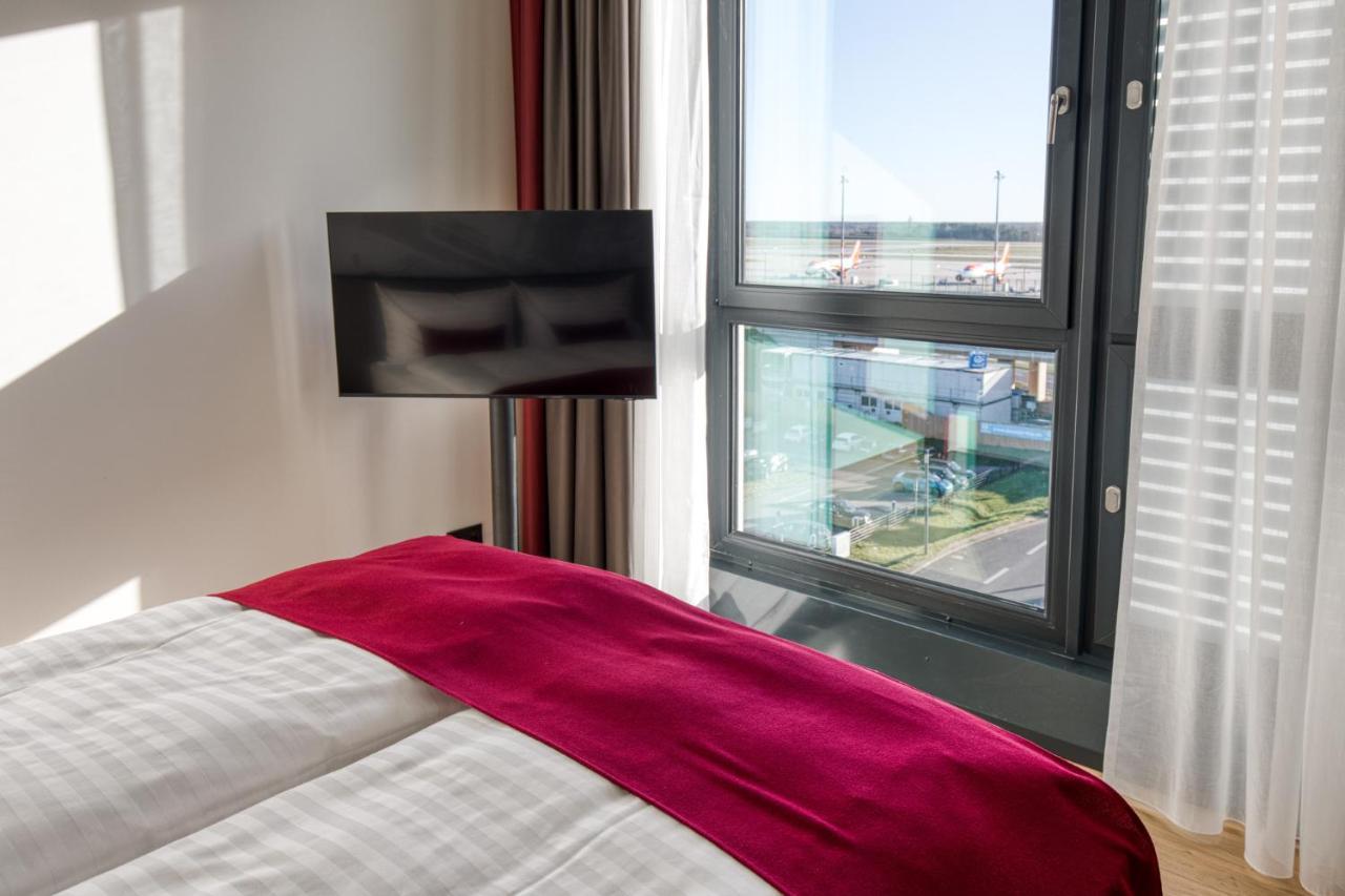 Intercityhotel Berlin Airport Ber Terminal 1 & 2 舍讷费尔德 外观 照片 The photo shows a modern hotel room with a large bed covered in white linens and a burgundy throw blanket. In the background, there is a large window that lets in natural light, offering a view of the outside. A flat-screen TV is mounted on the wall 