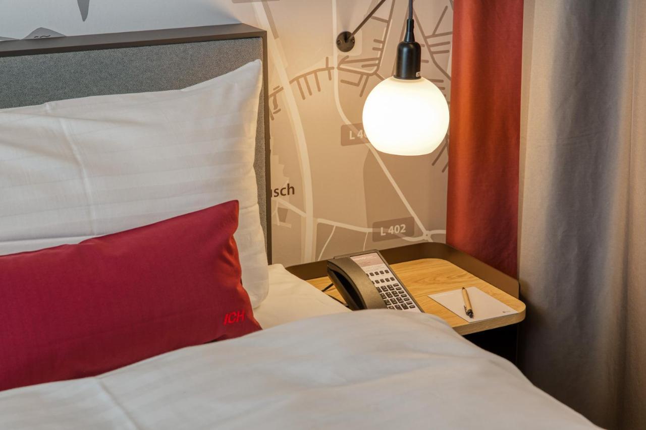 Intercityhotel Berlin Airport Ber Terminal 1 & 2 舍讷费尔德 外观 照片 The photo shows a cozy bedroom setup. There's a neatly made bed with white bedding and a red decorative pillow. Beside the bed, a small bedside table holds a telephone and a notebook or notepad, along with a pen. A round pendant light hangs above the