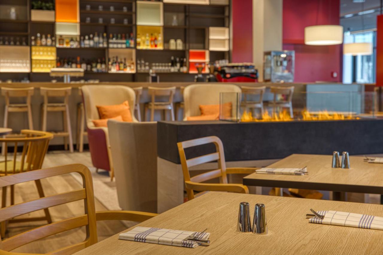 Intercityhotel Berlin Airport Ber Terminal 1 & 2 舍讷费尔德 外观 照片 The photo shows a modern restaurant or bar interior. In the foreground, there are wooden tables set with napkins and condiments. The seating includes wooden chairs and plush seating in various colors in the background. A stylish bar area is visible w